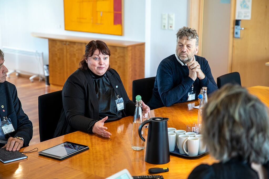 En gruppe mennesker satt rundt et konferansebord med kaffekopper; en kvinne snakker mens andre lytter oppmerksomt.
